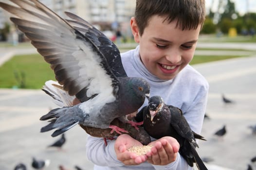 Close-up portrait of Caucasian smiling handsome adorable pre teen boy having fun during family outing, feeding birds from his hands in park square. The concept of kindness, love and care for animals