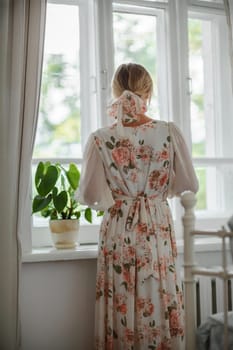 A woman wearing a cream dress with flowers stands by a window, lost in thought as she gazes at the scenery outside.