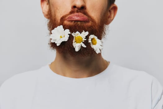 Portrait of a funny man in a white T-shirt with flowers daisies in his beard on a white isolated background, copy place. Holiday concept and congratulations. High quality photo