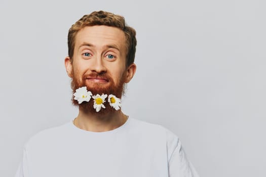 Portrait of a funny man in a white T-shirt with flowers daisies in his beard on a white isolated background, copy place. Holiday concept and congratulations. High quality photo