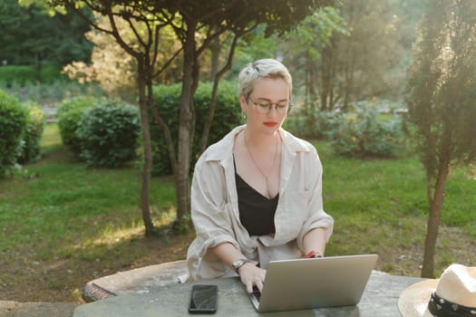 woman freelancer in glasses works at a computer at a white table in nature and spends her day productively.