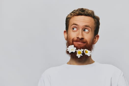 Portrait of a funny man in a white T-shirt with flowers daisies in his beard on a white isolated background, copy place. Holiday concept and congratulations. High quality photo