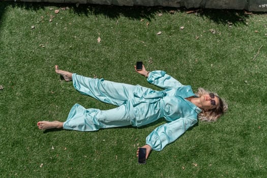 Women with blond hair in glasses and in a blue overalls lies on the summer fresh green grass with a phone in her hands. open image