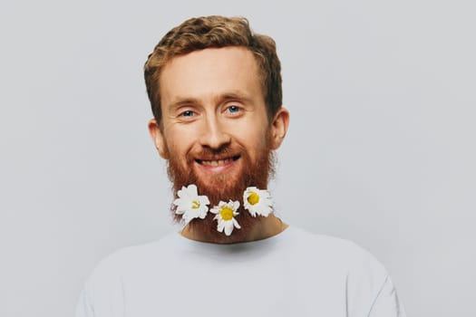 Portrait of a funny man in a white T-shirt with flowers daisies in his beard on a white isolated background, copy place. Holiday concept and congratulations. High quality photo