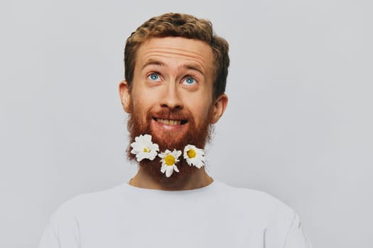Portrait of a funny man in a white T-shirt with flowers daisies in his beard on a white isolated background, copy place. Holiday concept and congratulations. High quality photo