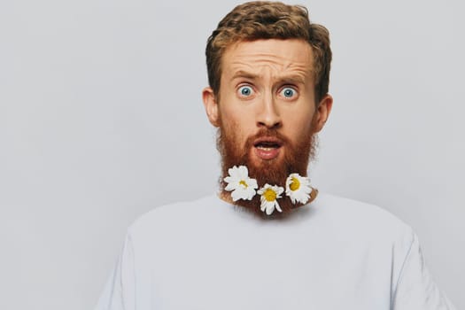 Portrait of a funny man in a white T-shirt with flowers daisies in his beard on a white isolated background, copy place. Holiday concept and congratulations. High quality photo