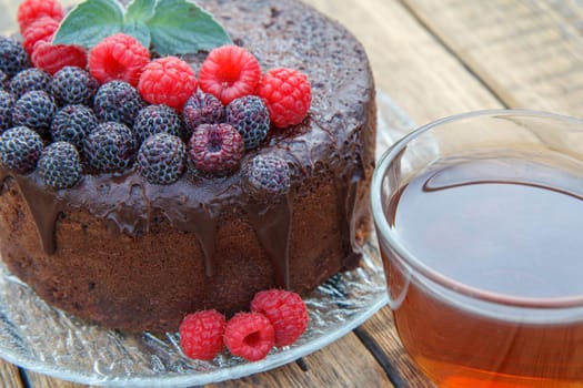 Homemade chocolate cake decorated with mint leaves, black and red raspberries on glass plate with cup of tea on wooden boards.