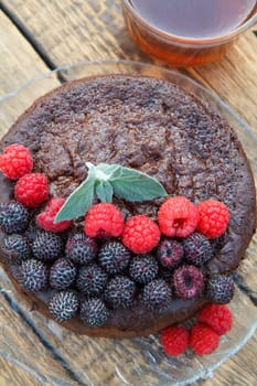 Homemade chocolate cake decorated with mint leaves, black and red raspberries on glass plate with cup of tea on wooden boards. Top view.