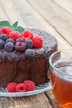 Homemade chocolate cake decorated with mint leaves, black and red raspberries on glass plate with cup of tea on wooden boards.