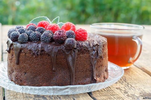 Homemade chocolate cake decorated with mint leaves, black and red raspberries on glass plate with cup of tea on wooden boards.