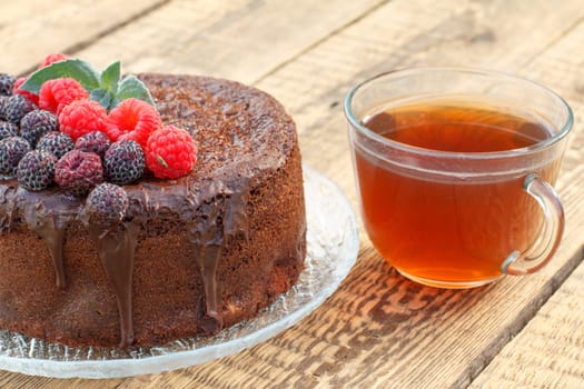 Homemade chocolate cake decorated with mint leaves, black and red raspberries on glass plate with cup of tea on wooden boards.