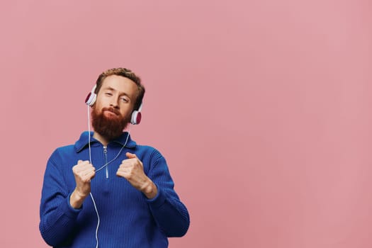 Portrait of a redheaded man wearing headphones smiling and dancing, listening to music on a pink background. A hipster with a beard. High quality photo