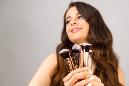 Coquettish young girl holding cosmetic brushes in her hands on the grey background with copy space.