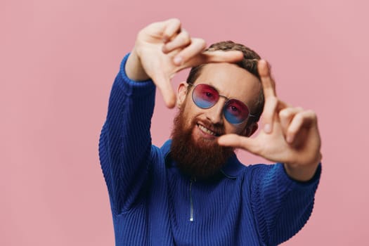 Portrait of a redheaded man wearing sunglasses smiling and dancing, listening to music on a pink background. Hipster with a beard, happiness finger pointing. High quality photo