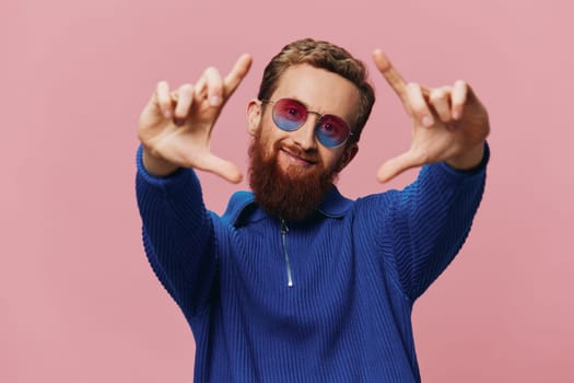 Portrait of a redheaded man wearing sunglasses smiling and dancing, listening to music on a pink background. Hipster with a beard, happiness finger pointing. High quality photo