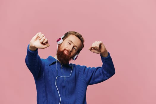 Portrait of a redheaded man wearing headphones smiling and dancing, listening to music on a pink background. A hipster with a beard. High quality photo