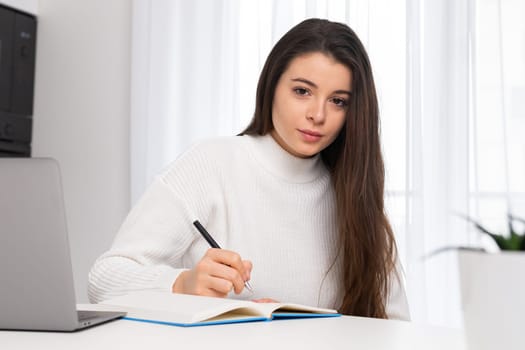 Portrait of a female college student sitting on the desk and writing in the textbook.