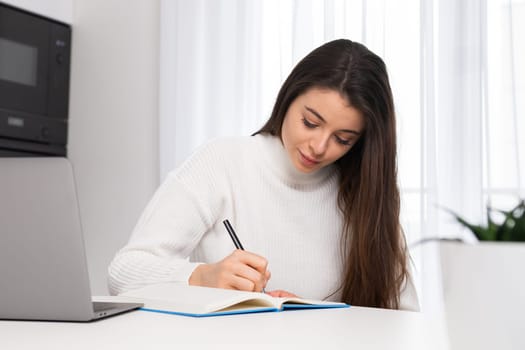 Beautiful student watching the webinar and making notes sitting in the kitchen