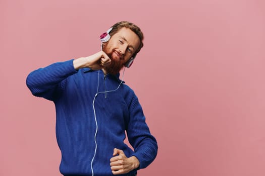 Portrait of a redheaded man wearing headphones smiling and dancing, listening to music on a pink background. A hipster with a beard. High quality photo