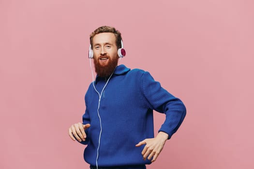 Portrait of a redheaded man wearing headphones smiling and dancing, listening to music on a pink background. A hipster with a beard. High quality photo