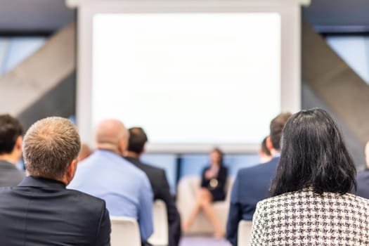 Business and entrepreneurship symposium. Female speaker giving a talk at business meeting. Audience in conference hall. Rear view of unrecognized participant in audience.