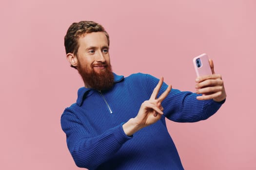 Portrait of a redheaded man with phone in hand taking selfies and photos on his phone with a smile on a pink background, blogger. High quality photo