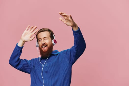 Portrait of a redheaded man wearing headphones smiling and dancing, listening to music on a pink background. A hipster with a beard. High quality photo