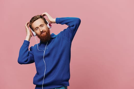 Portrait of a redheaded man wearing headphones smiling and dancing, listening to music on a pink background. A hipster with a beard. High quality photo