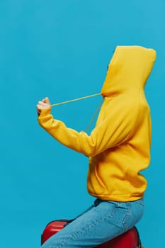 Smile woman sitting on a suitcase in a yellow hoodie, blue jeans and glasses on a blue background, packing for a trip. High quality photo