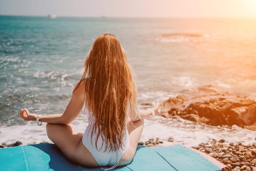 Young woman in swimsuit with long hair practicing stretching outdoors on yoga mat by the sea on a sunny day. Women's yoga fitness pilates routine. Healthy lifestyle, harmony and meditation concept.