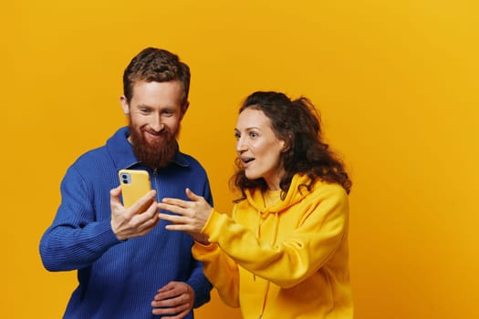 Man and woman couple smiling merrily with phone in hand social media viewing photos and videos, on yellow background, symbols signs and hand gestures, family freelancers. High quality photo