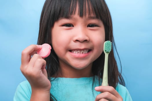 Smiling cute little girl holding toothbrush and sweets isolated on blue background. Kid training oral hygiene and Unhealth eat. Tooth decay prevention or dental care concept.