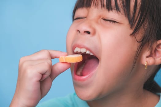 Smiling cute little girl eating sweet gelatin with sugar added isolated on blue background. Children eat sugary sweets, causing loss teeth or tooth decay and unhealthy oral care.