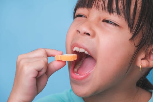 Smiling cute little girl eating sweet gelatin with sugar added isolated on blue background. Children eat sugary sweets, causing loss teeth or tooth decay and unhealthy oral care.