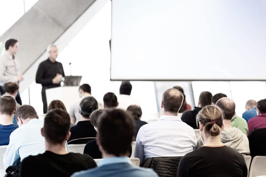 Men giving presentation in lecture hall. Male speeker having talk at public event. Participants listening to lecture. Rear view, focus on people in audience.