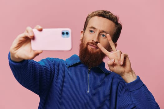 Portrait of a redheaded man with phone in hand taking selfies and photos on his phone with a smile on a pink background, blogger. High quality photo