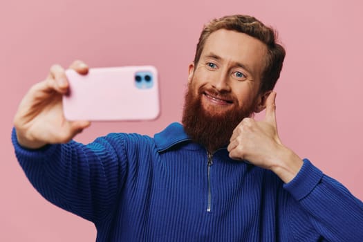 Portrait of a redheaded man with phone in hand taking selfies and photos on his phone with a smile on a pink background, blogger. High quality photo