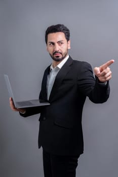 Successful businessman in black suit with innovative tech concept, finger pointing at camera and holding laptop and smiling with excitement on copyspace background for promotion or ad. Fervent