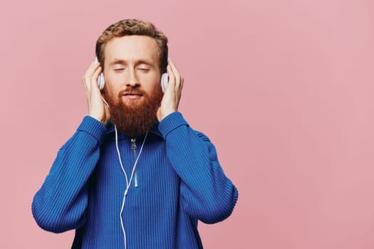 Portrait of a redheaded man wearing headphones smiling and dancing, listening to music on a pink background. A hipster with a beard. High quality photo