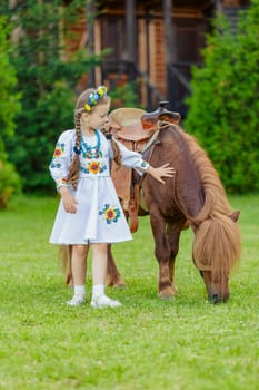 young girl in Ukrainian national dress strokes a pony that grazes on the lawn