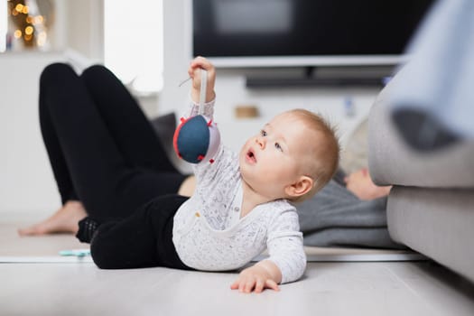 Happy family moments. Mother lying comfortably on children's mat watching and suppervising her baby boy playinghis in living room. Positive human emotions, feelings, joy.