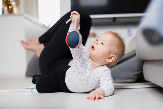 Happy family moments. Mother lying comfortably on children's mat watching and suppervising her baby boy playinghis in living room. Positive human emotions, feelings, joy.