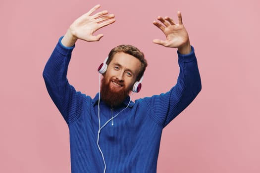 Portrait of a redheaded man wearing headphones smiling and dancing, listening to music on a pink background. A hipster with a beard. High quality photo