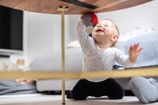 Cute infant baby boy playing with hanging ball, crawling and standing up by living room table at home. Baby activity and play center for early infant development. Baby playing at home