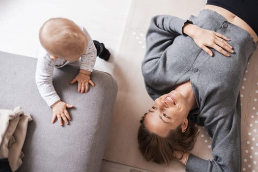 Happy family moments. Mother lying comfortably on children's mat playing with her baby boy watching and suppervising his first steps. Positive human emotions, feelings, joy