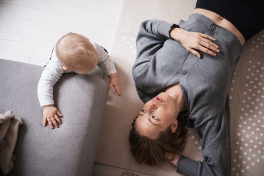 Happy family moments. Mother lying comfortably on children's mat playing with her baby boy watching and suppervising his first steps. Positive human emotions, feelings, joy