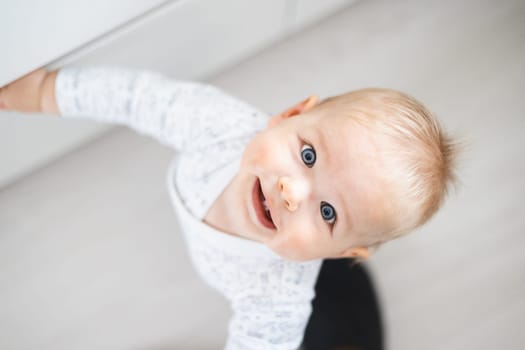 top down view of cheerful baby boy infant taking first steps holding to kitchen drawer at home. Cute baby boy learning to walk.