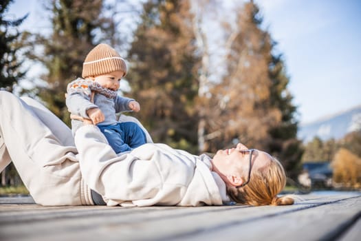 Happy family. Young mother playing with her baby boy infant oudoors on sunny autumn day. Portrait of mom and little son on wooden platform by lake. Positive human emotions, feelings, joy