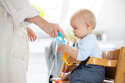 Happy infant sitting at dining table and playing with his toy in traditional scandinavian designer wooden high chair in modern bright atic home superwised by his mother.