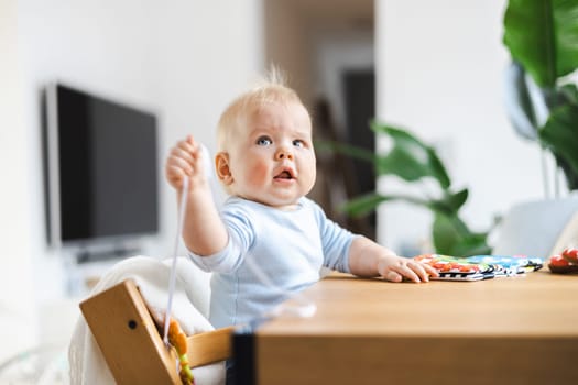 Happy infant sitting at dining table and playing with his toy in traditional scandinavian designer wooden high chair in modern bright atic home. Cute baby playing with toys.